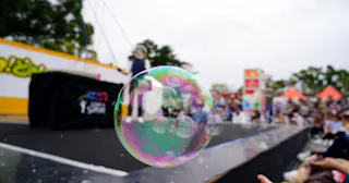 A large, iridescent soap bubble floats in sharp focus at an outdoor event, with people, colorful tents, and a stage blurred in the background.
