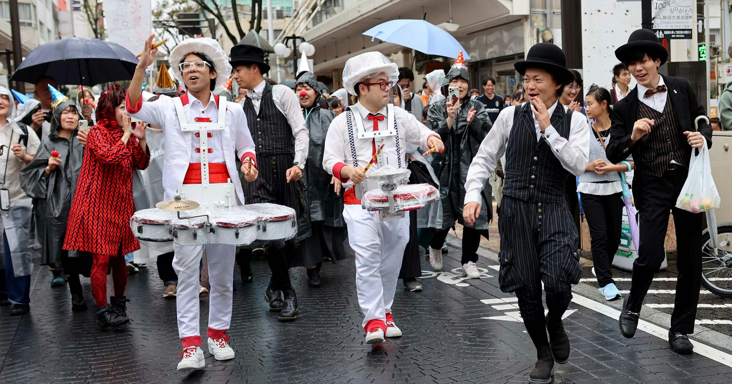 A lively street parade with musicians in playful costumes playing drums, surrounded by smiling people in raincoats and hats. Some carry umbrellas as they walk on a wet city street. The mood is festive and joyful.