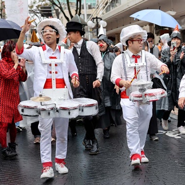 Daidogei World Cup A lively street parade with musicians in playful costumes playing drums, surrounded by smiling people in raincoats and hats. Some carry umbrellas as they walk on a wet city street. The mood is festive and joyful.