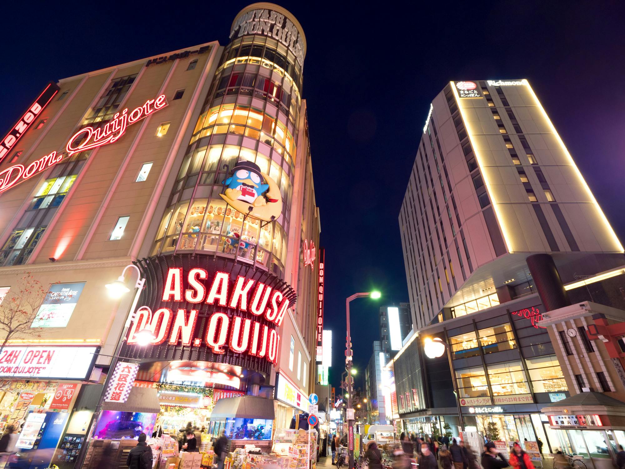 A busy Tokyo street at night featuring the brightly lit Don Quijote store in Asakusa, with neon signs and tall buildings, as people walk along the sidewalks.