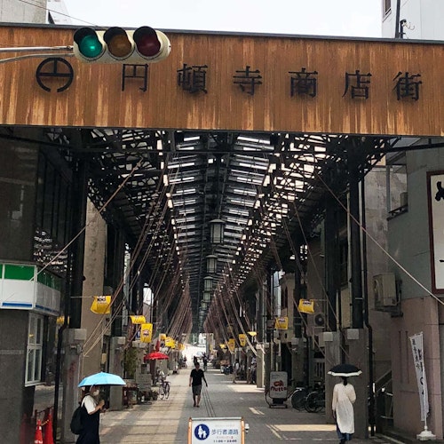 Endoji Shotengai Shopping Street A covered shopping street entrance in Japan, with a large wooden sign overhead. People walk under umbrellas on the street, and small shops line both sides under the tall, metal-framed canopy.