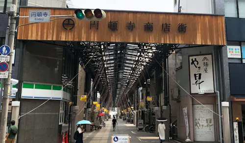 A covered shopping street entrance in Japan, with a large wooden sign overhead. People walk under umbrellas on the street, and small shops line both sides under the tall, metal-framed canopy.