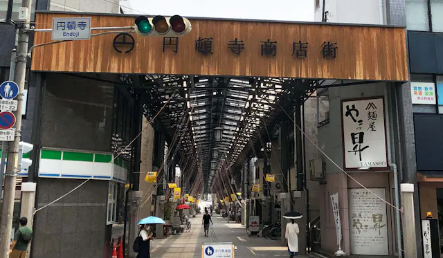 A covered shopping street entrance in Japan, with a large wooden sign overhead. People walk under umbrellas on the street, and small shops line both sides under the tall, metal-framed canopy.