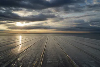 Sunset over a body of water, with dramatic clouds in the sky and sunlight reflecting off a smooth, glass-like surface that stretches toward the horizon.