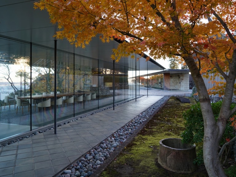 Enoura Observatory Modern glass-walled building with a long dining table inside, surrounded by autumn trees with orange leaves and a stone path outside. The scene is tranquil and reflective.
