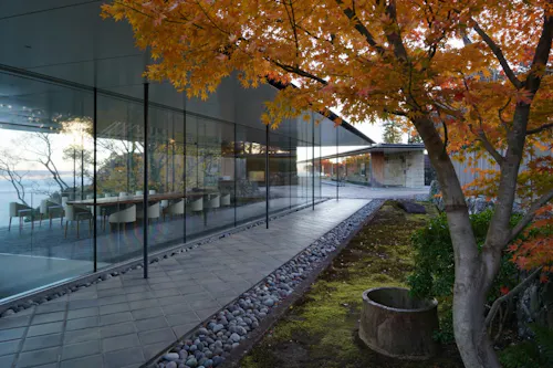 Modern glass-walled building with a long dining table inside, surrounded by autumn trees with orange leaves and a stone path outside. The scene is tranquil and reflective.