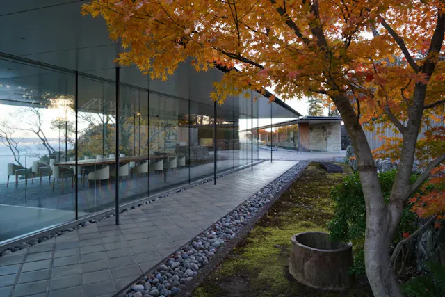 Modern glass-walled building with a long dining table inside, surrounded by autumn trees with orange leaves and a stone path outside. The scene is tranquil and reflective.