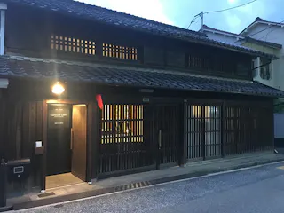 A traditional Japanese wooden building with sliding lattice doors and tiled roof, softly lit at dusk. A sign by the entrance reads "Enpukuji Gallery." The street in front is empty.