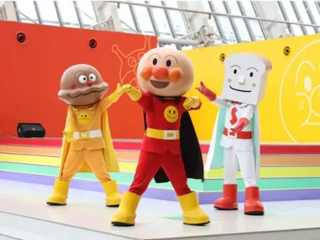 Three people in colorful superhero costumes pose on a bright stage. One is dressed as a burger, another as a round-faced character with red cheeks, and the third as a slice of bread. The backdrop is vibrant and playful.