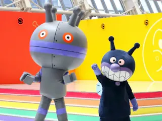 Two people in colorful robot and monster costumes, one gray with antennas and the other dark blue with a big toothy grin, stand on a rainbow-striped stage with a bright yellow and orange background.