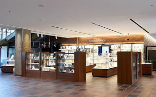 A well-lit museum shop with wooden shelves and glass displays filled with books, souvenirs, and various items, located in a spacious lobby with tiled flooring.