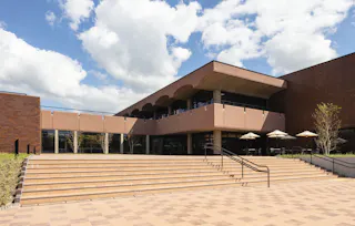 A modern building with large windows and a shaded terrace, featuring wide tile steps leading up to the entrance. Outdoor tables with umbrellas are visible on the right, and the sky is partly cloudy.