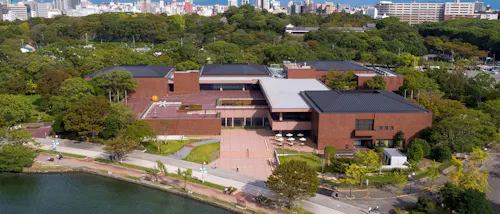 Aerial view of a modern brick building surrounded by trees and greenery, with a courtyard and outdoor seating; a cityscape is visible in the background. A pond borders the walkway in the foreground.