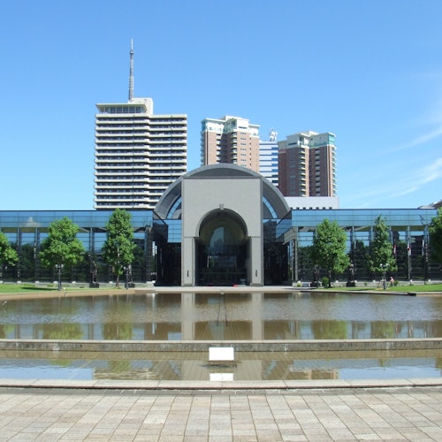Fukuoka City Museum A modern glass building with an arched entrance stands behind a reflective pond, surrounded by green trees, with tall city buildings visible in the background under a clear blue sky.