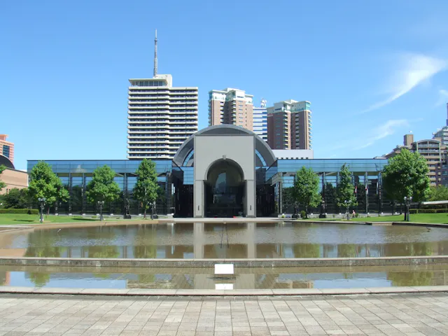 A modern glass building with an arched entrance stands behind a reflective pond, surrounded by green trees, with tall city buildings visible in the background under a clear blue sky.