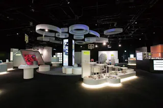 A modern science museum exhibit features interactive displays on white circular platforms, illuminated by soft lighting, with large abstract rings suspended from the dark ceiling above.
