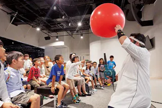 A group of excited children sit on benches watching an adult in a white coat and headband holding up a large red balloon inside a brightly lit indoor space.