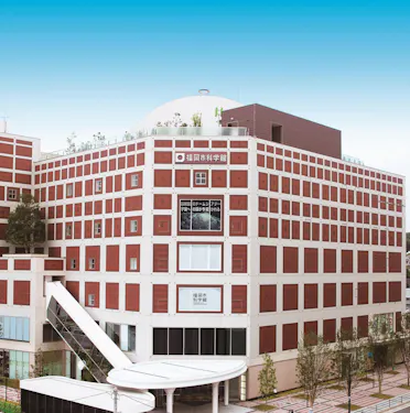 A modern, multi-story building with a red and white grid pattern exterior, large windows, and a rooftop garden. A sky bridge connects to another structure. The sky is clear and blue.