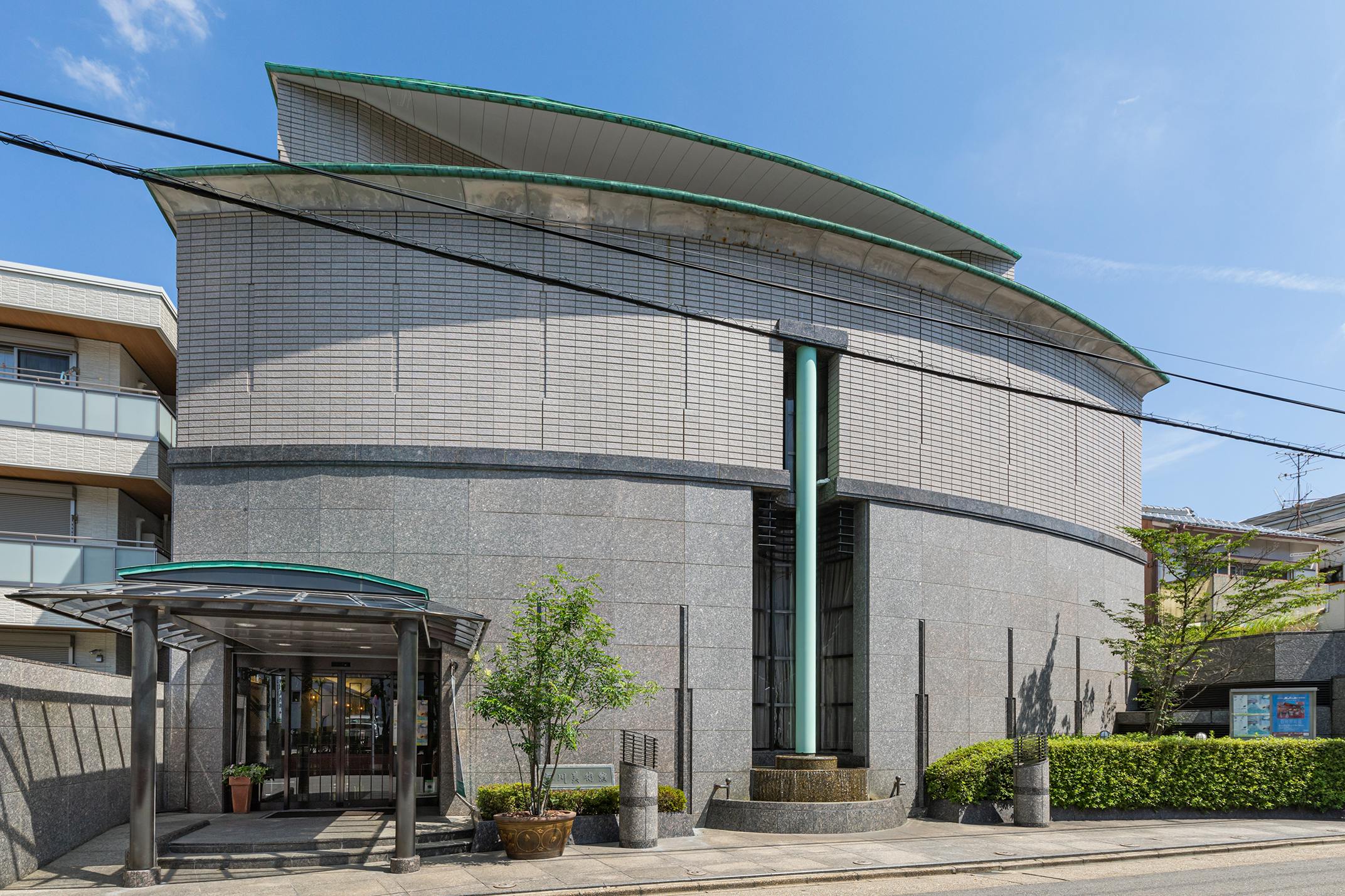 A modern building with curved gray walls, a green-trimmed roof, and vertical glass feature, with a covered entrance, potted plants, and trees along the sidewalk, under a clear blue sky.