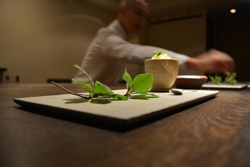 A close-up of a rectangular plate with green leaves on a wooden table. In the background, a person in a white shirt reaches toward another dish, which is slightly out of focus.