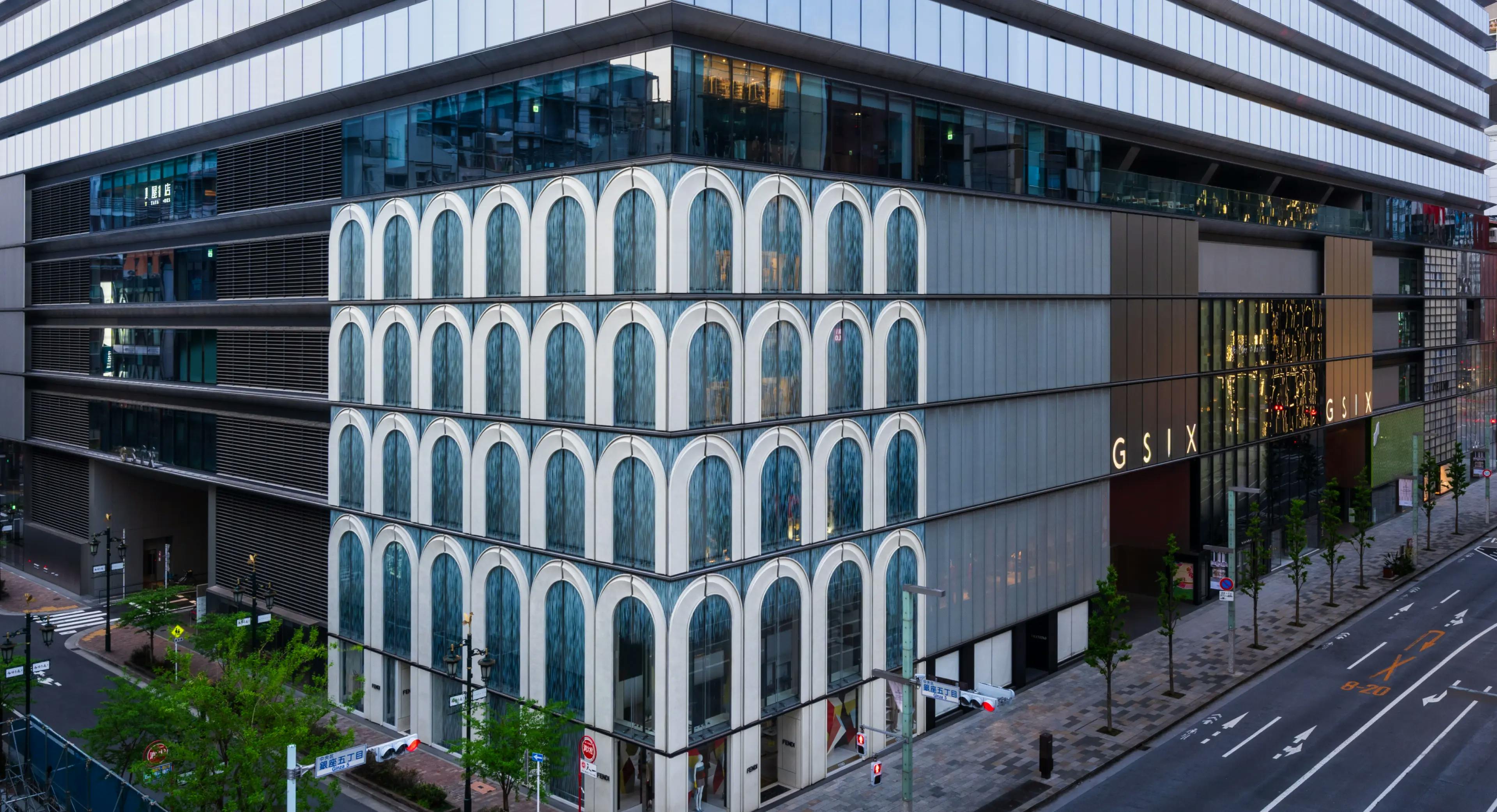 A modern city building with a white façade featuring multiple arched windows, located at a street corner. The adjacent building displays the letters “GSIX” in gold on a dark background. The streets around are empty.