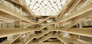 A modern atrium with geometric lighting on the ceiling, multiple floors, glass railings, and zigzagging staircases connecting the levels in a symmetrical design.