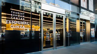 The entrance of a modern Tourist Service Center called Terminal Ginza, with large glass doors and illuminated signage. Inside, shelves with brochures and products are visible, along with reflections of city buildings in the glass.