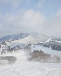 Snow-covered mountains and trees under a partly cloudy sky, with rolling hills and forested areas visible in the distance, creating a serene winter landscape.