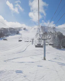 A ski lift ascends a snowy mountain under a blue sky with scattered clouds. Freshly groomed ski trails and snow-covered trees line both sides of the wide, white slope.