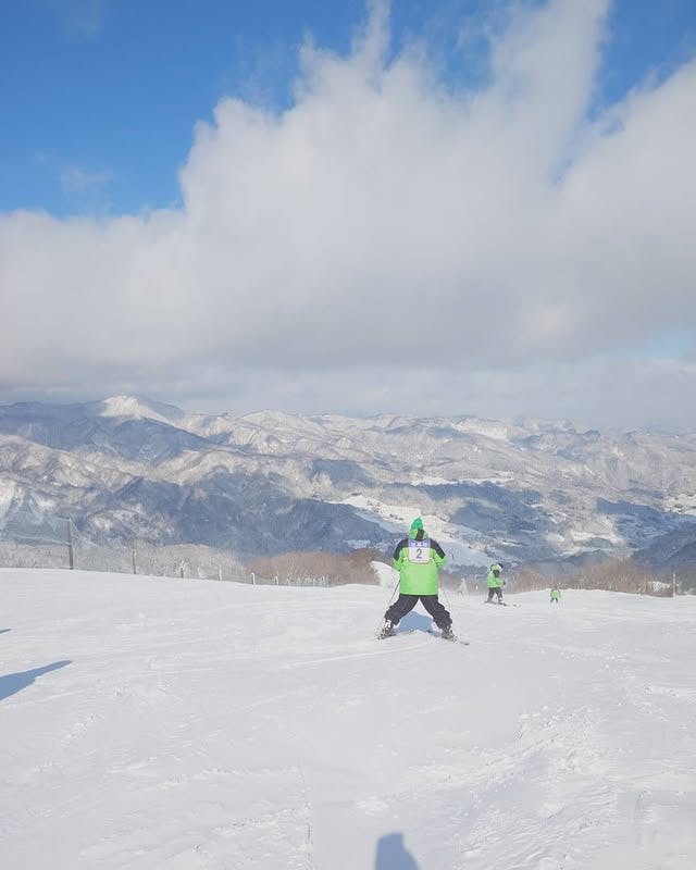 Skiers in green jackets descend a snowy slope under a blue sky with clouds, with snow-covered mountains and valleys visible in the distance.