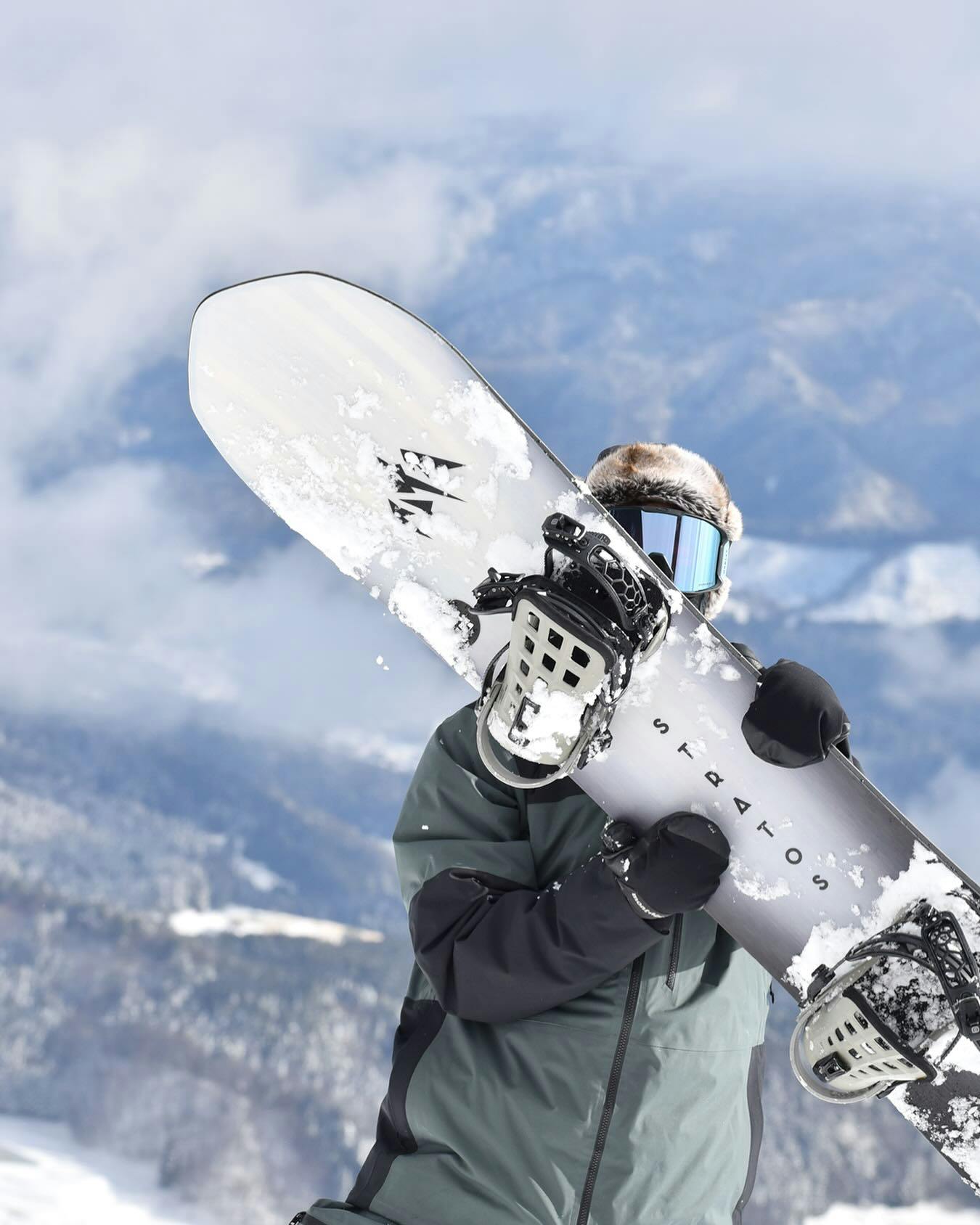 A snowboarder in winter gear and goggles holds a snow-covered snowboard upright in front of their body with snowy mountains and cloudy sky in the background.