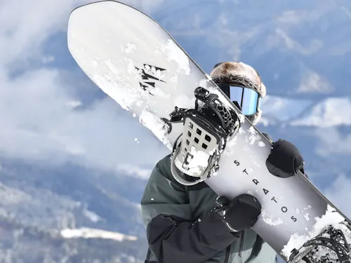 Hachi Kita Kogen Ski Resort A snowboarder in winter gear and goggles holds a snow-covered snowboard upright in front of their body with snowy mountains and cloudy sky in the background.