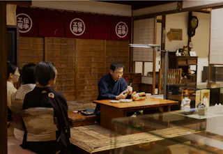 A craftsman sits at a wooden table in a traditional Japanese workshop, demonstrating pottery techniques to visitors dressed in kimonos. Various ceramics are displayed around the room.