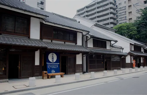 Traditional Japanese wooden buildings with tiled roofs line a quiet street, contrasting with modern apartment buildings in the background. A blue sign in Japanese hangs by the entrance of one building.