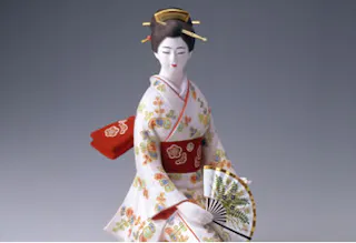 A traditional Japanese doll dressed in a floral-patterned kimono and red obi, holding a decorative folding fan. The doll has a serene expression and wears a traditional hair ornament.