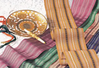 A gold incense holder with a stick rests on white stones beside colorful, striped textiles in shades of purple, green, gold, and blue, arranged in gentle folds.