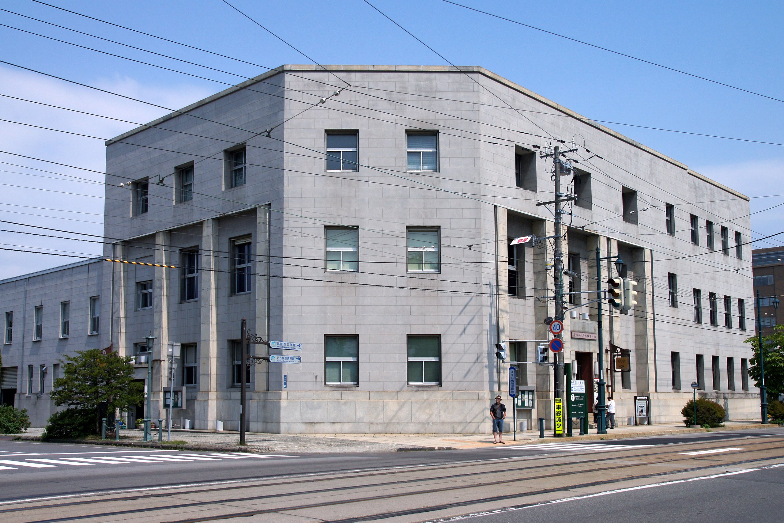 A gray, three-story, rectangular building with many windows stands on a street corner with traffic lights, power lines, and two people waiting at the crosswalk on a clear day.