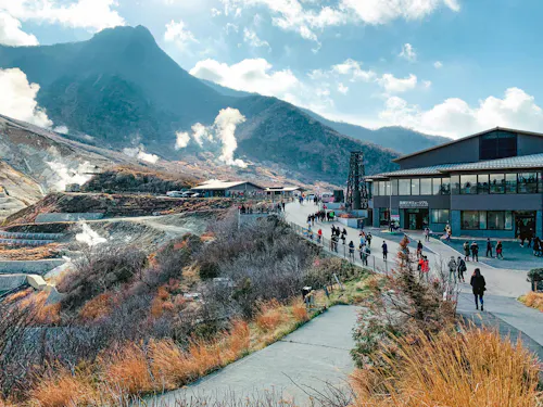 Hakone Geo Museum People walk along a path near a modern building in a mountainous area with steam rising from the ground. The sky is partly cloudy, and the landscape features brown grass and shrubs.