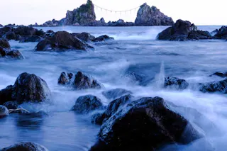 Rocky shoreline with crashing waves in the foreground and two large, rocky islets connected by a sacred rope in the distance, under a cloudy sky.