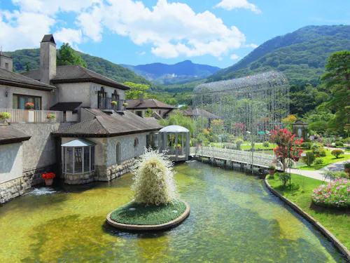 Hakone Glass no Mori Venetian Glass Museum A scenic view of an elegant stone building beside a pond with a small central fountain, surrounded by lush gardens, trees, and mountains under a bright blue sky with clouds.