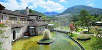 A scenic view of an elegant stone building beside a pond with a small central fountain, surrounded by lush gardens, trees, and mountains under a bright blue sky with clouds.