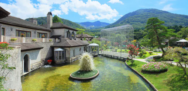 A scenic view of an elegant stone building beside a pond with a small central fountain, surrounded by lush gardens, trees, and mountains under a bright blue sky with clouds.