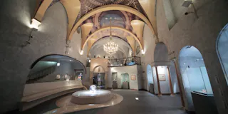 Elegant museum interior with vaulted ceilings, arched doorways, a large chandelier, and a central glass display case. Artwork and exhibits line the walls, and a staircase leads to an upper balcony level.