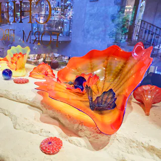 A display of colorful blown glass sculptures, including large, wavy orange and red pieces and smaller round and shell-shaped objects, arranged on a stone surface in front of a window.