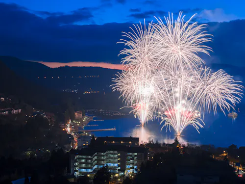Hakone Summer Festival Week Bright white fireworks explode over a lake at dusk, illuminating the water and the surrounding town buildings, with mountains and a dark blue sky in the background.