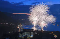 Bright white fireworks explode over a lake at dusk, illuminating the water and the surrounding town buildings, with mountains and a dark blue sky in the background.