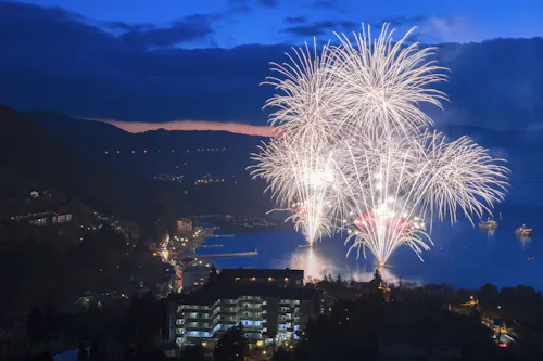 Bright white fireworks explode over a lake at dusk, illuminating the water and the surrounding town buildings, with mountains and a dark blue sky in the background.