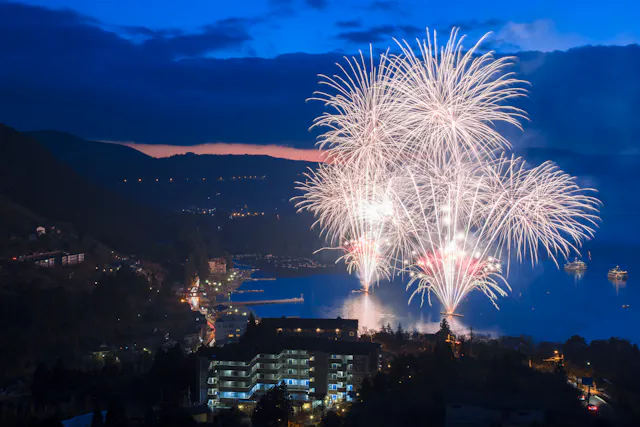 Bright white fireworks explode over a lake at dusk, illuminating the water and the surrounding town buildings, with mountains and a dark blue sky in the background.