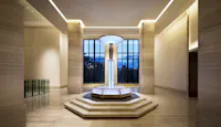 A modern, spacious lobby features a central, illuminated glass fountain on a tiered hexagonal base, with large floor-to-ceiling windows and beige marble walls and floors.