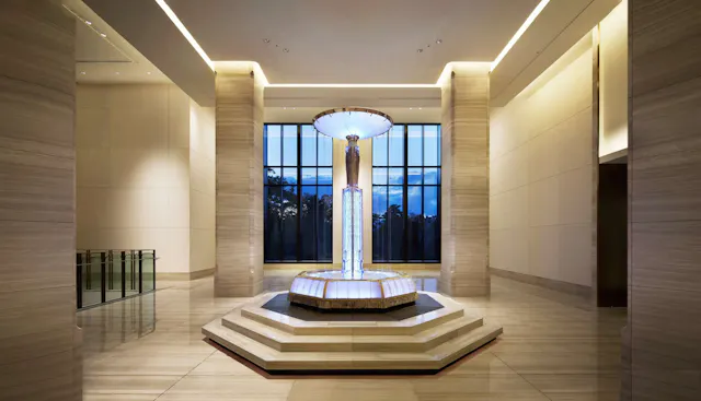 A modern, spacious lobby features a central, illuminated glass fountain on a tiered hexagonal base, with large floor-to-ceiling windows and beige marble walls and floors.